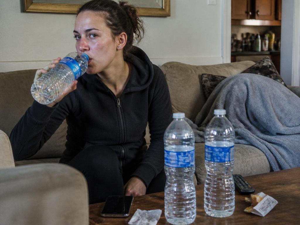 A woman slumped on a couch at home drinking from an oversized one-litre water bottle, more big bottles on the cluttered coffee table