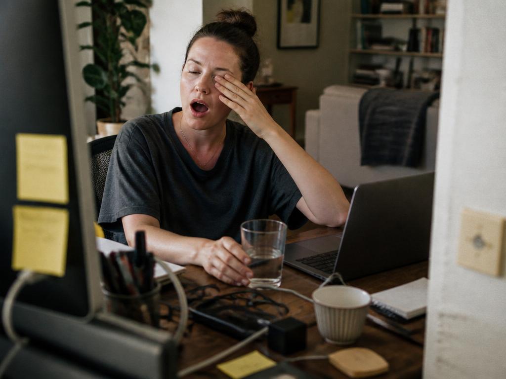 A tired woman yawning and rubbing her eye at a messy home-office desk in the afternoon, a half-full glass of water beside her laptop