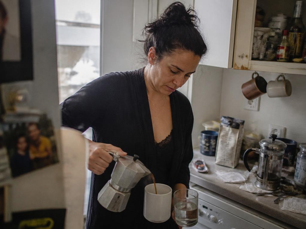 A sleepy woman in a cramped kitchen pouring fresh coffee from a moka pot into a mug, a half-finished glass of water beside it