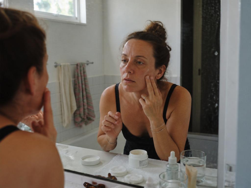 A woman leaning close to a bathroom mirror patting moisturizer onto her bare, unretouched cheek, a glass of water on the cluttered counter