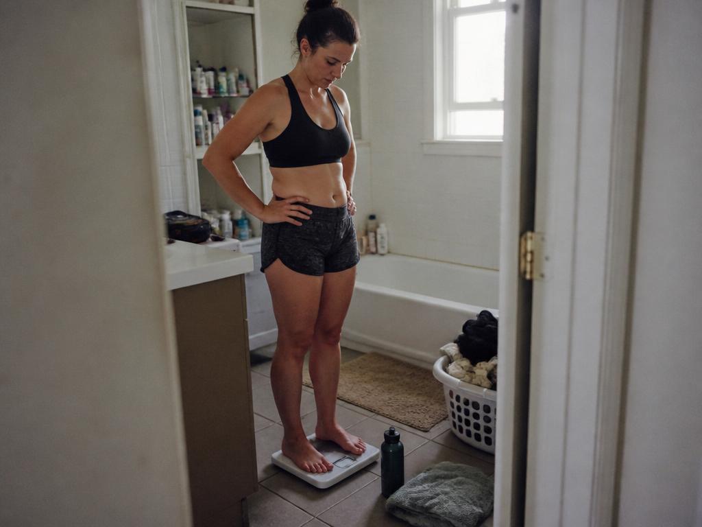 A sweaty woman in workout clothes standing on a bathroom scale just after exercise, looking down at the number, a towel and water bottle on the floor