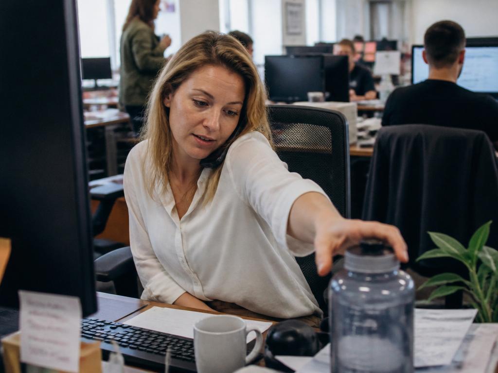 A woman at a cluttered open-plan office desk reaching for a large refillable water bottle while still looking at her monitors, phone at her ear