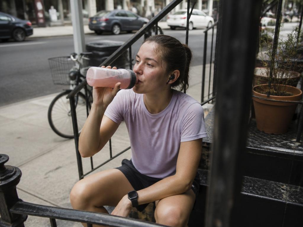 A flushed woman sitting on a city stoop just after a run, tipping a sports water bottle to her mouth and catching her breath
